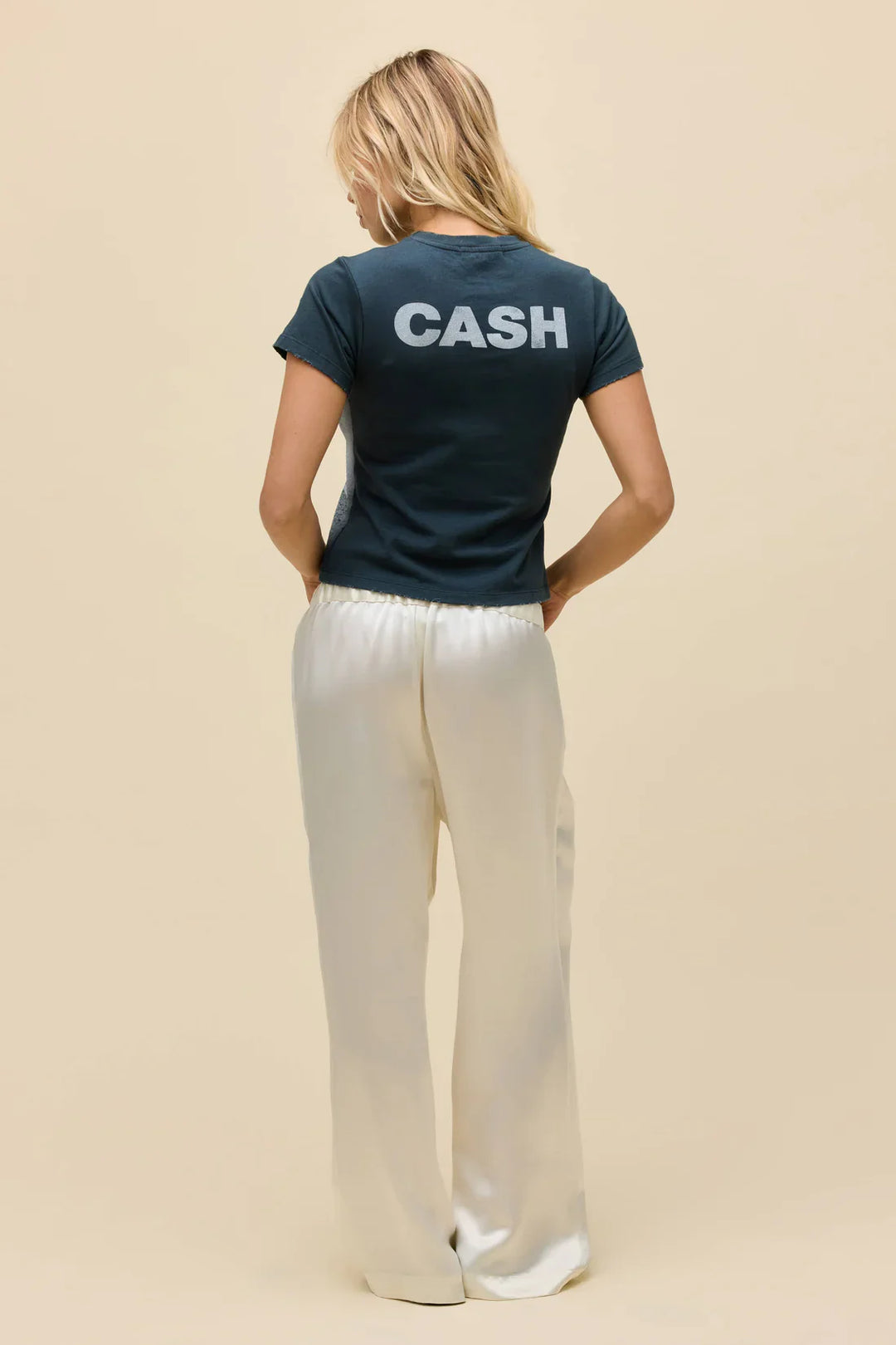 A woman faces away in the Zertz JOHNNY CASH SILHOUETTE VNTG TEE, featuring bold “CASH” lettering on the back, paired with loose white pants against a plain beige background, channeling country legend Johnny Cash’s iconic style.