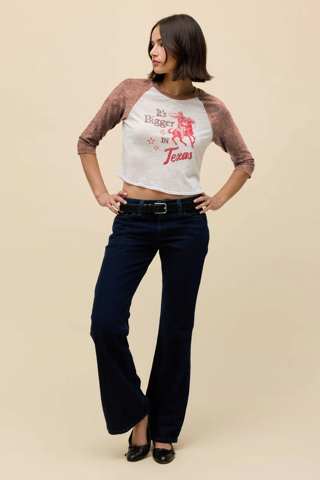 A woman with shoulder-length dark hair wears the Zertz ITS BIGGER IN TX BURNOUT RAGLAN TEE, featuring a cowboy graphic, paired with dark blue jeans, a black belt, and black shoes. She stands confidently with hands on her hips.