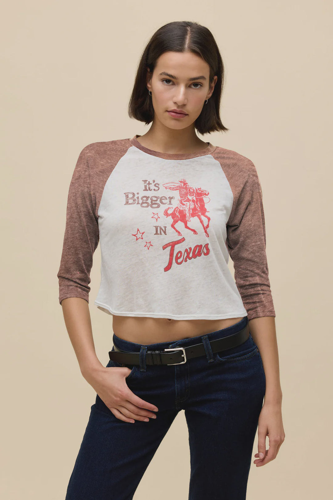 A woman with short brown hair wears the Zertz ITS BIGGER IN TX BURNOUT RAGLAN TEE featuring a bold cowboy graphic, styled with dark jeans and a black belt, standing in front of a beige background.