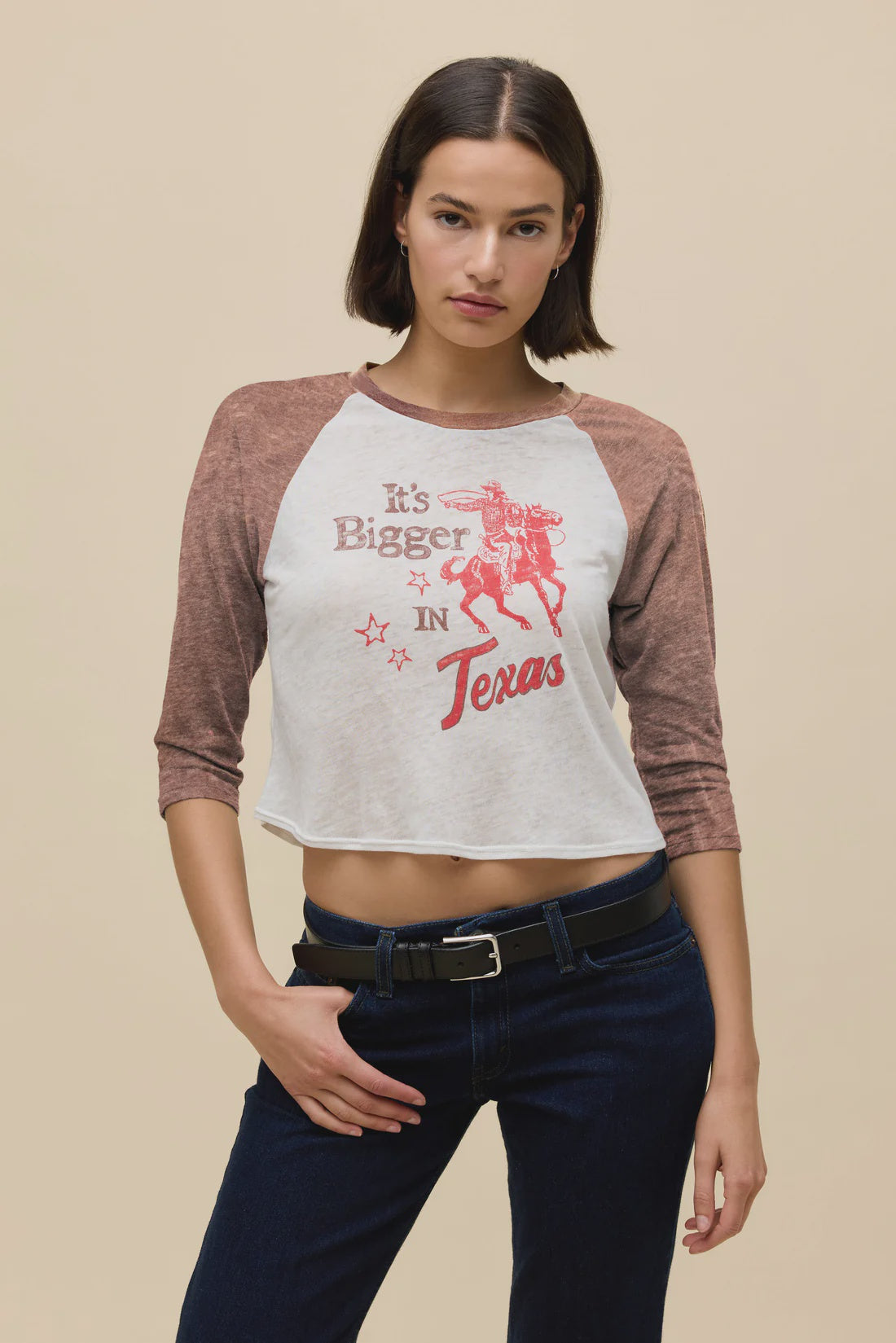 A woman with short brown hair wears the Zertz ITS BIGGER IN TX BURNOUT RAGLAN TEE featuring a bold cowboy graphic, styled with dark jeans and a black belt, standing in front of a beige background.
