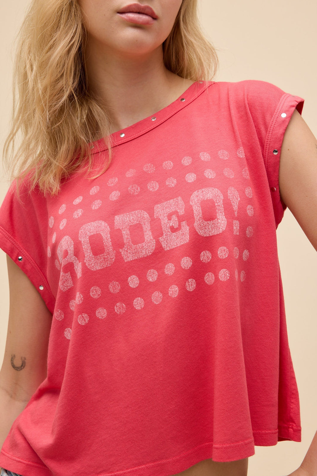 A woman models the Zertz RODEO! MUSCLE TANK, a loose pink-red top with silver studs and “RODEO!” in crackled white ink among dots. The image captures her upper body and part of her face.