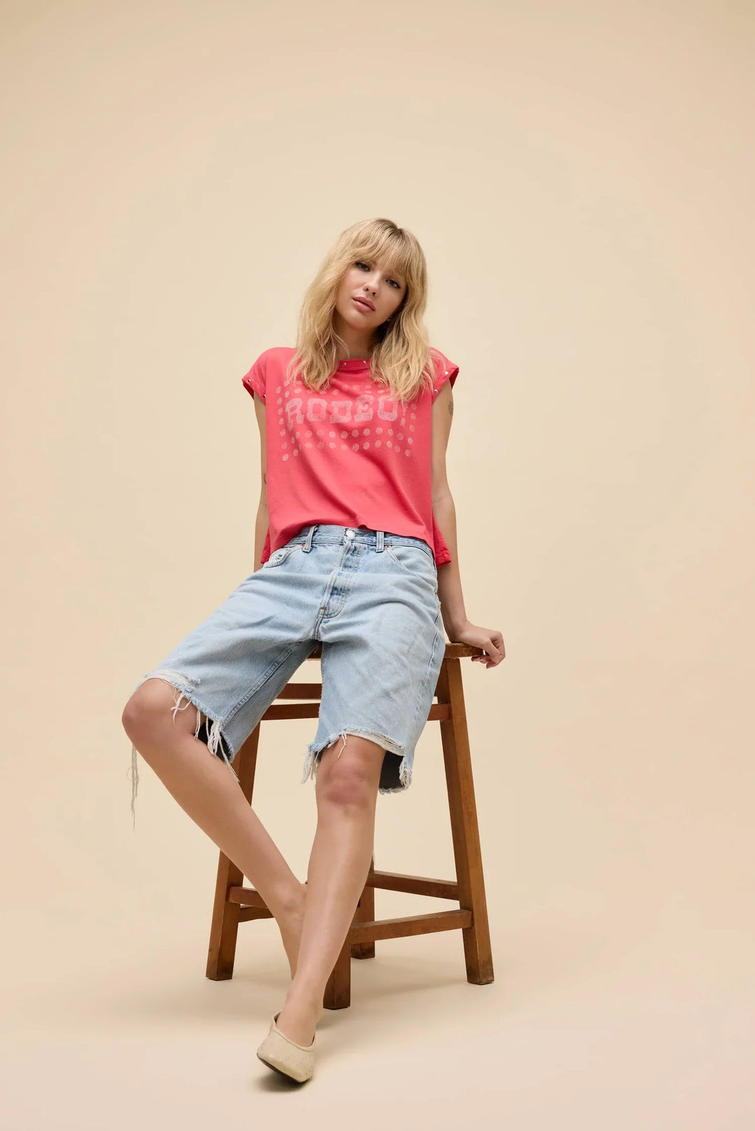 A blonde-haired person sits barefoot on a wooden stool against a plain beige background, wearing the Zertz RODEO! MUSCLE TANK in bright pink with crackle ink and light blue knee-length denim shorts.