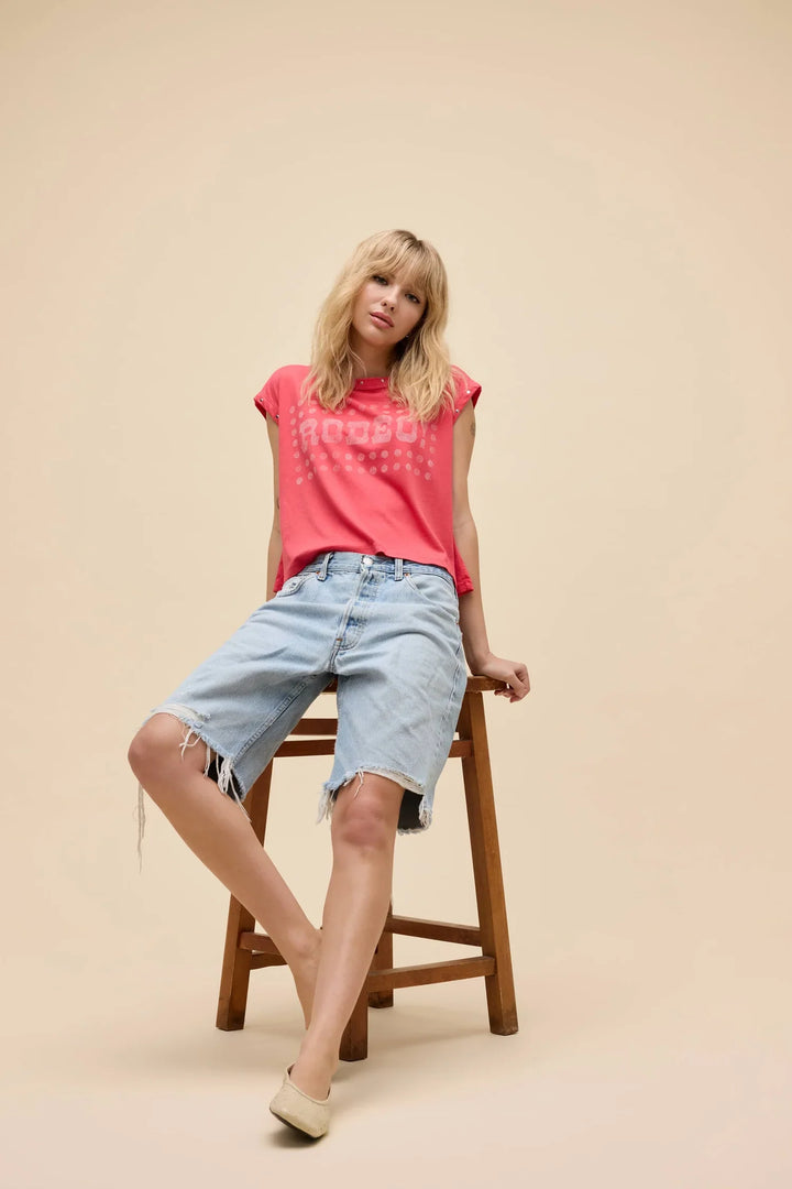 A blonde-haired person sits barefoot on a wooden stool against a plain beige background, wearing the Zertz RODEO! MUSCLE TANK in bright pink with crackle ink and light blue knee-length denim shorts.