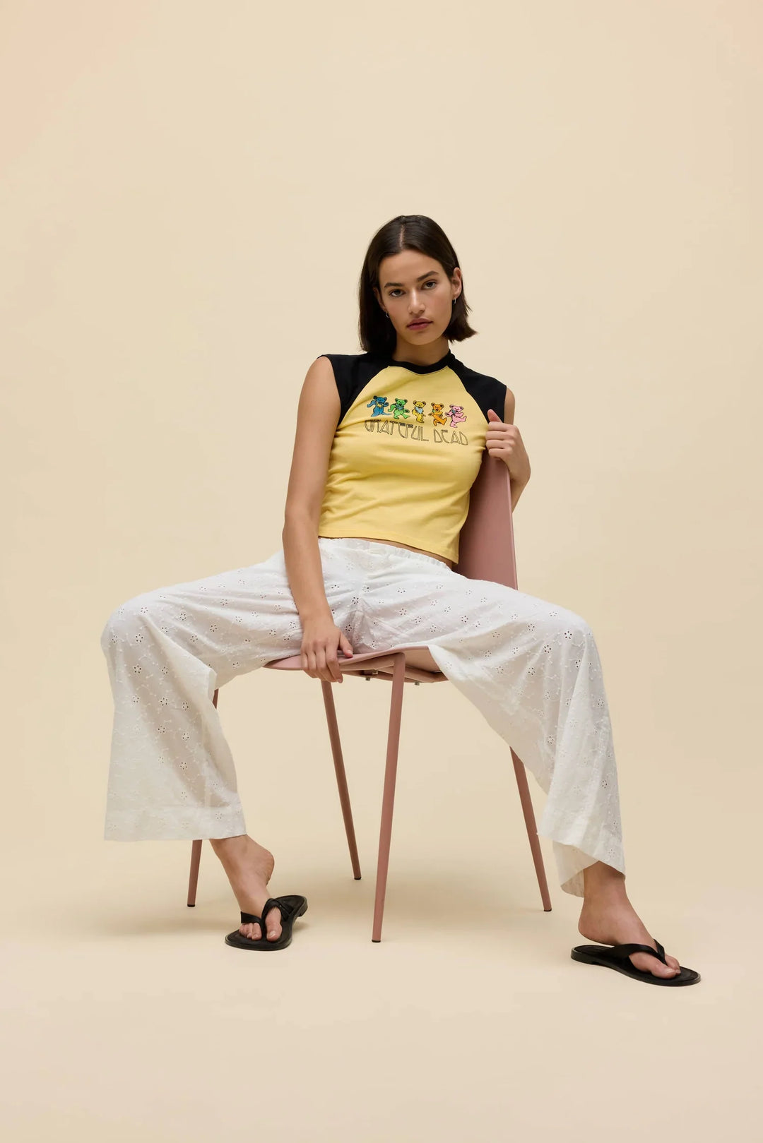 A woman sits on a pink chair with legs apart, wearing the yellow Zertz Grateful Dead Bears Raglan Tee, white eyelet pants, and black sandals against a beige background.
