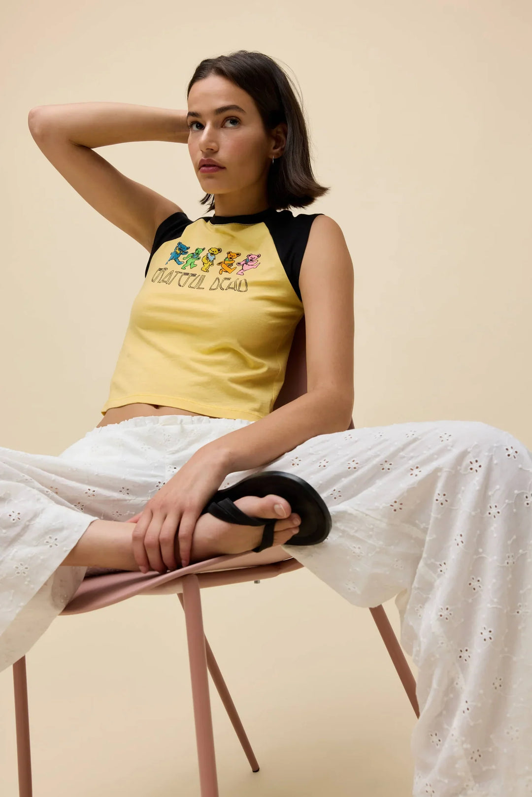 A woman sits on a chair with legs apart, wearing the Zertz GREATFUL DEAD BEARS RGLAN TEE, white eyelet pants, and black sandals. She rests one arm on her head and looks into the camera against a beige background.