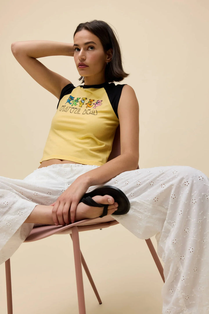 A woman sits on a chair with legs apart, wearing the Zertz GREATFUL DEAD BEARS RGLAN TEE, white eyelet pants, and black sandals. She rests one arm on her head and looks into the camera against a beige background.