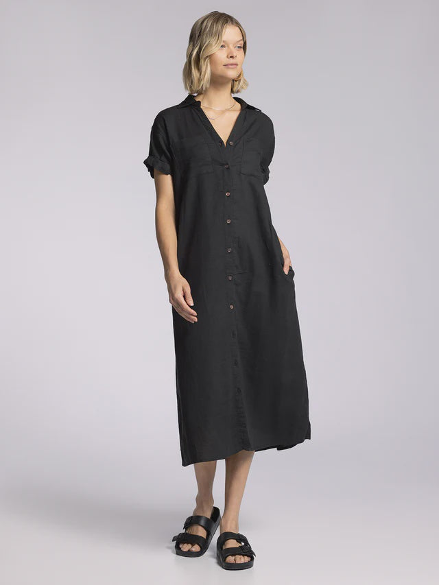 A woman stands against a plain background wearing the Zertz PARIS DRESS, a loose-fitting black midi dress with a collared neckline, short sleeves, button-down front, and pockets. She pairs it with black slide sandals.