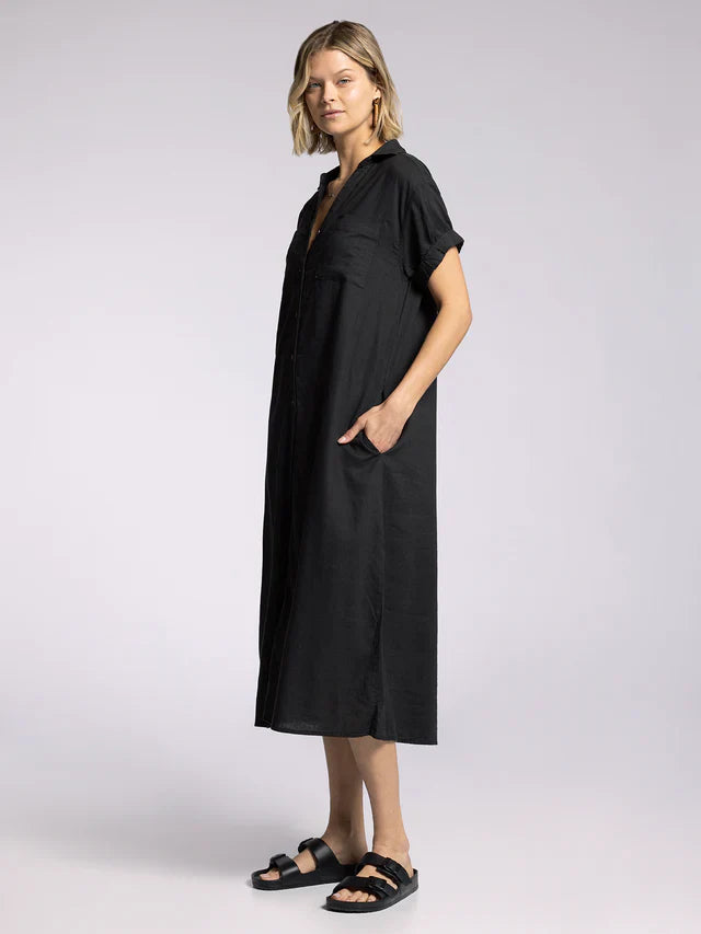 A woman stands sideways in the Zertz PARIS DRESS, a loose black midi with short sleeves, collared neckline, buttons, and side pockets, paired with black sandals against a plain light background.
