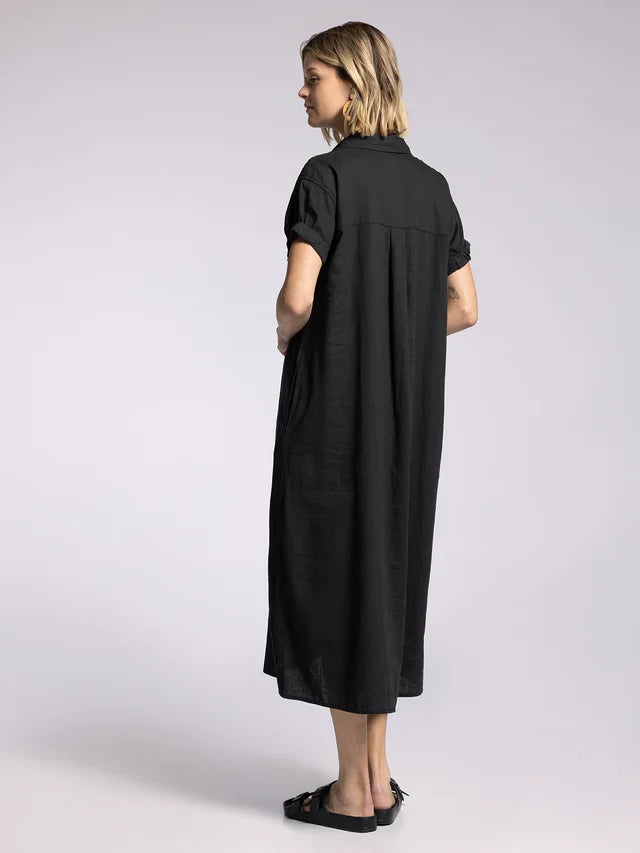 A person stands sideways, looking left, wearing the Zertz PARIS DRESS—a loose-fit black midi dress with a collared neckline and short sleeves—paired with black sandals against a plain light gray background.