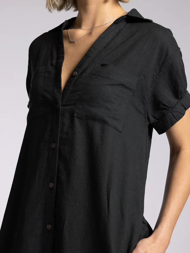 A person wearing the Zertz PARIS DRESS, a loose-fitting black dress with a collared neckline and short sleeves, slightly unbuttoned at the top and styled with a delicate silver necklace.