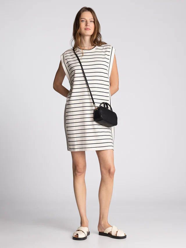 A woman with light brown hair and a neutral expression models the Zertz AVELIN DRESS, a sleeveless white mini dress with black horizontal stripes, paired with white sandals and a small black crossbody purse, in a studio setting.