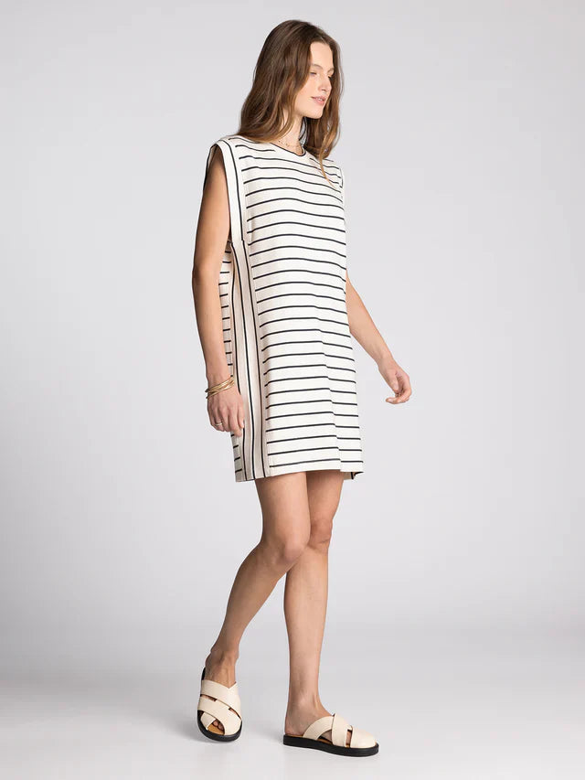 A woman models the AVELIN DRESS by Zertz, a cream, knee-length sleeveless shirt dress with black horizontal stripes. She pairs it with light sandals and gold bracelets, set against a plain light gray background.