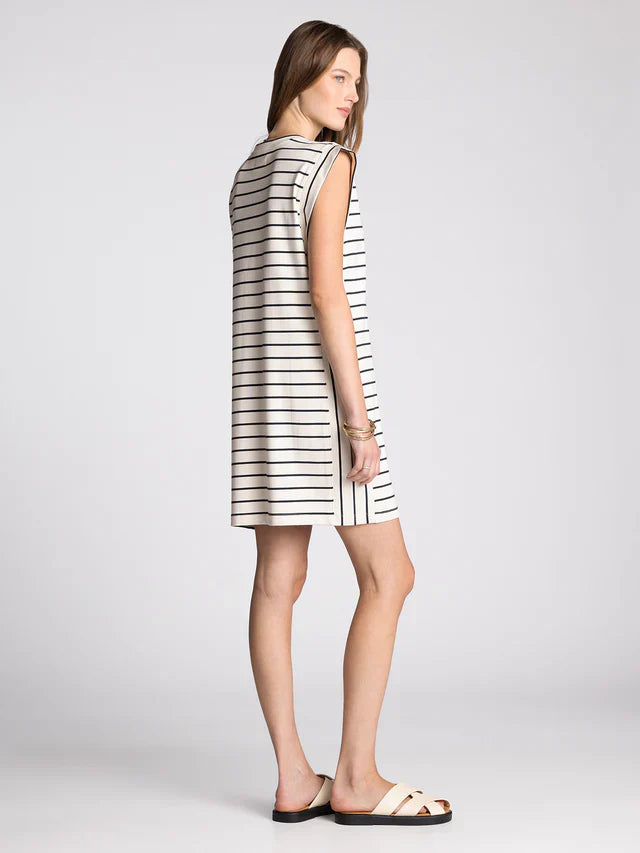 A woman with light brown hair poses sideways against a light gray background wearing the Zertz AVELIN DRESS—a relaxed fit, sleeveless, knee-length white dress with black horizontal stripes—paired with white sandals.