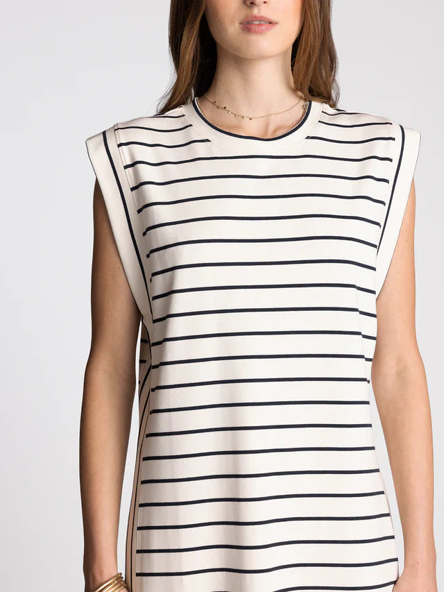 A woman wears the AVELIN DRESS by Zertz, a sleeveless white dress with black horizontal stripes and padded shoulders. With a relaxed fit, she adds a simple chain necklace. The photo is cropped from shoulders to mid-thigh.