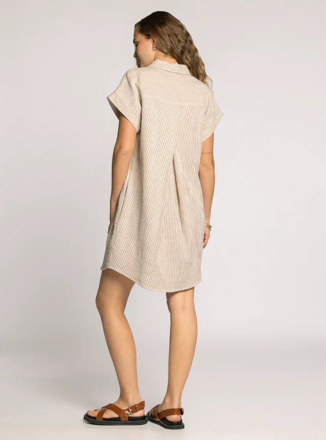 A woman with wavy hair stands facing away on a light background, wearing Zertz’s CRESCENT DRESS—a short-sleeved, knee-length shirt dress in beige and white stripes with a collar—paired with brown sandals.