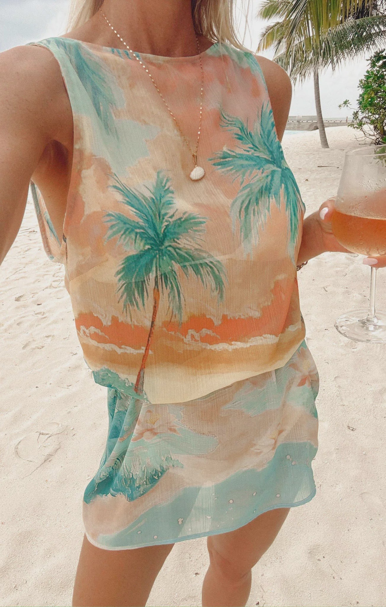 On a sandy beach, a woman wears the Zertz KIKI MINI DRESS SUNSET COVE—a lightweight chiffon mini dress with pastel palm prints—holding rosé wine. A seashell necklace and palm trees adorn the background.