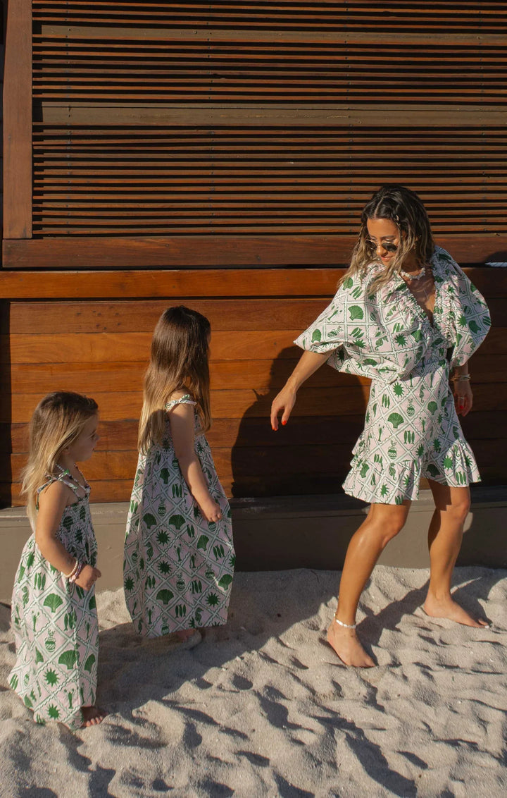 A woman and two young girls stand on sand before a wooden wall, all wearing matching Zertz HADLEY MINI DRESS outfits in green-and-white coastal collage print with puff sleeves; the woman appears to be playing or dancing with them.