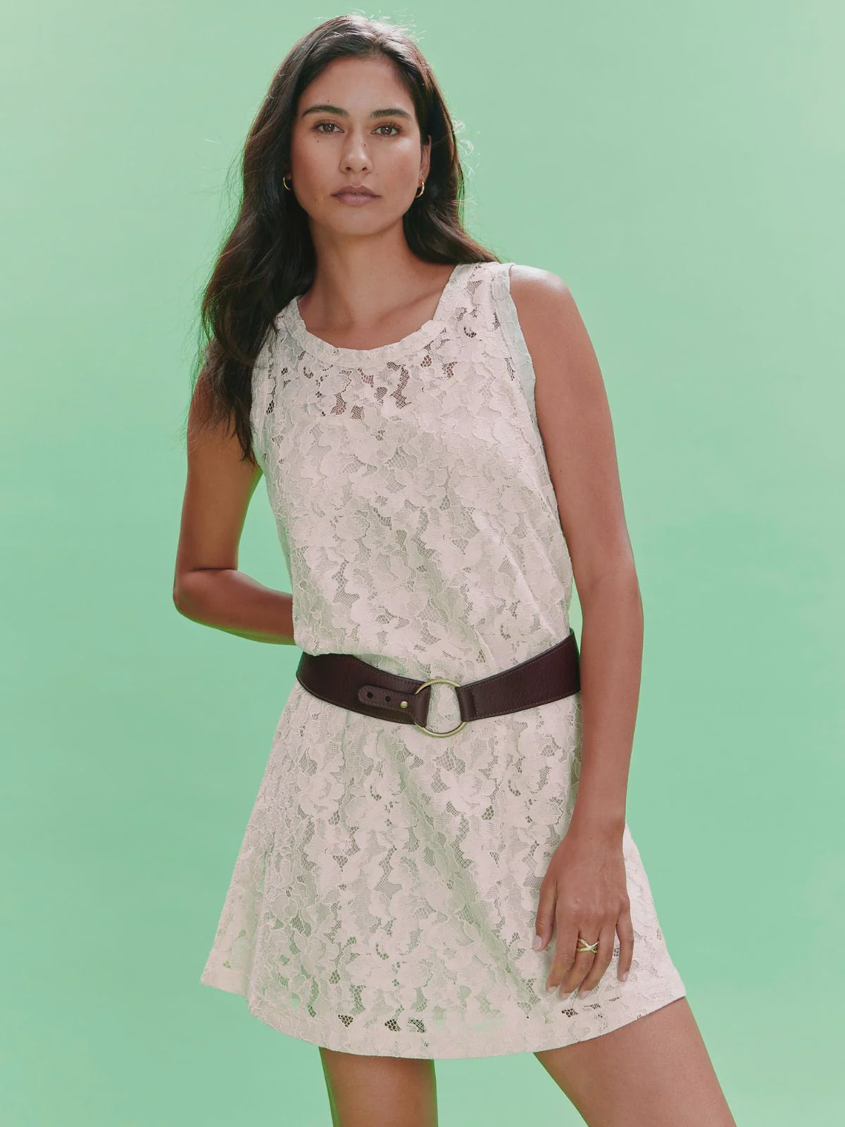 Against a plain light green background, a woman with long dark hair and a neutral expression wears the Zertz SUNDAY BEST LACE TANK DRESS, a chic sleeveless mini dress with a dark brown belt and detachable slip.