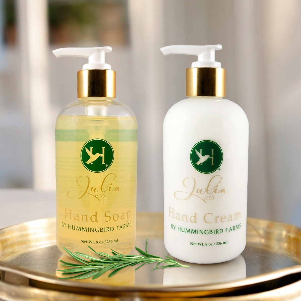 The Julia 1933 Hand Soap & Cream Duo by Hummingbird Farms, featuring yellow aloe vera hand soap and white whipped hand cream, sits on a gold tray with a sprig of rosemary in front.