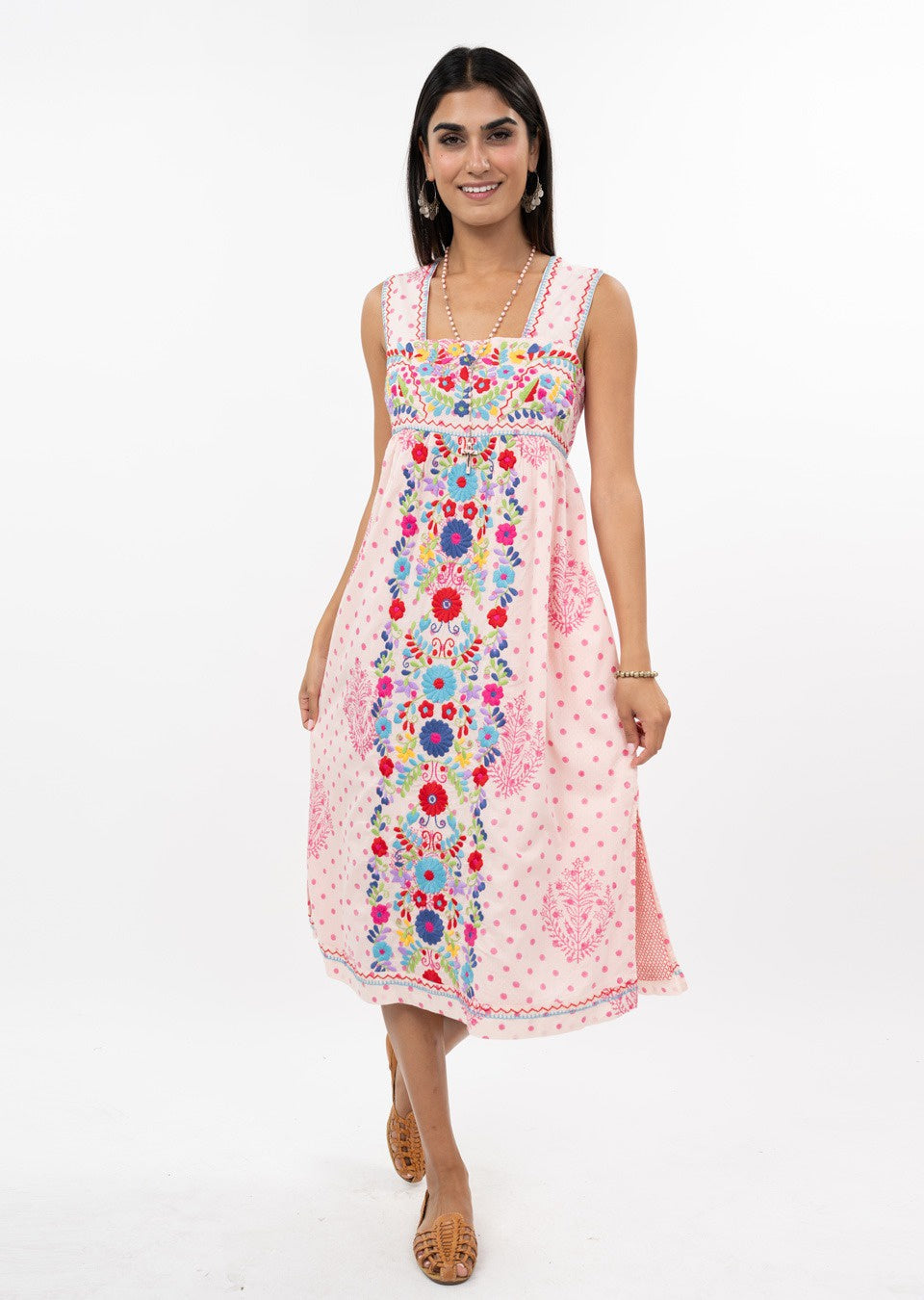A woman in the VALNTINA EMBD FLOWER MIDI DRESS by Zertz—sleeveless, knee-length, pink with floral embroidery—smiles against a white background. Styled with gold jewelry and sandals, this look is perfect for special occasions.