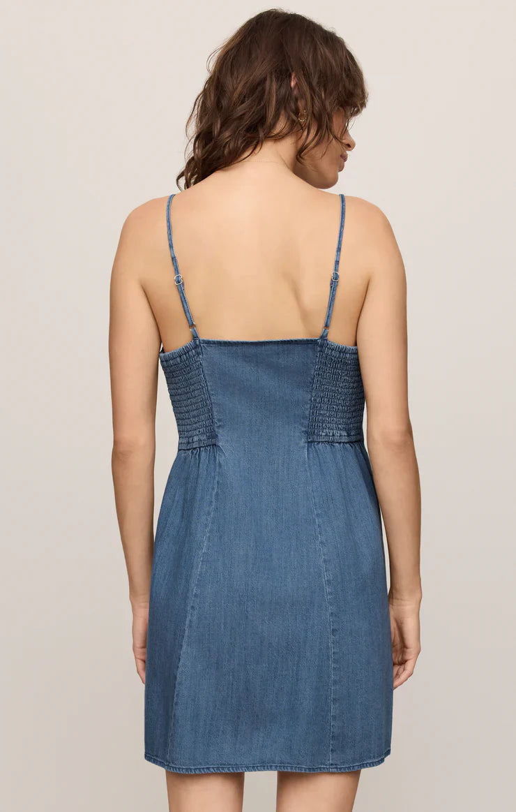 A woman with wavy brown hair faces away, wearing the Zertz ANGEL EYES DENIM MINI DRESS MEDIUM INDIGO—a blue, cotton-blend mini dress with thin shoulder straps and a smocked back—set against a light plain background.