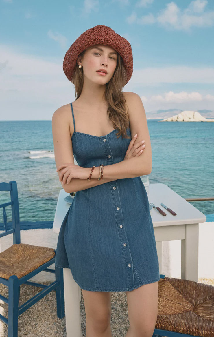 A woman wearing the Zertz ANGEL EYES DENIM MINI DRESS MEDIUM INDIGO stands with arms crossed by a white table near the sea, with blue chairs and a clear sky in the background.