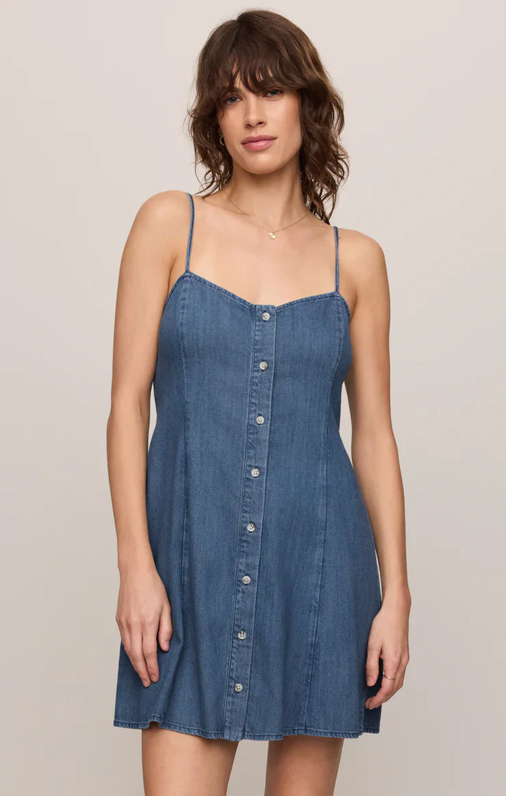 A woman with wavy brown hair models the Zertz ANGEL EYES DENIM MINI DRESS in medium indigo, featuring thin straps and a smocked bodice, against a plain light background.