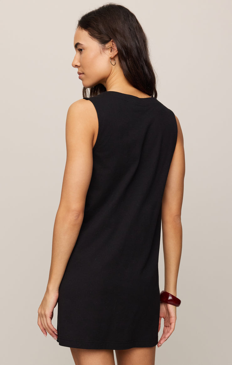 A woman with long dark hair stands facing away, wearing the Zertz SOLACE MINI DRESS TRUE BLACK, a sleeveless black v-neck mini dress. She accessorizes with a brown bracelet on her right wrist against a light beige background.