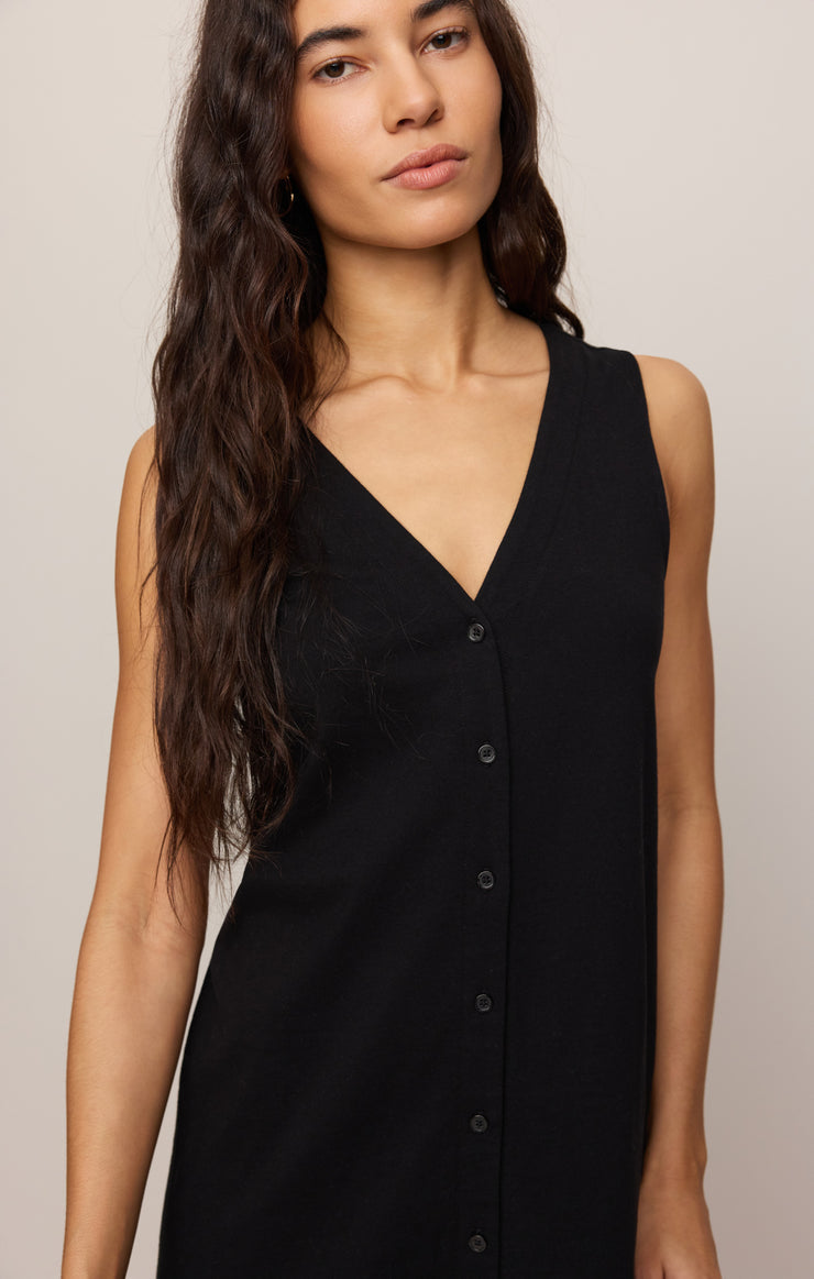 A woman with long, wavy dark hair models the Zertz SOLACE MINI DRESS TRUE BLACK—a sleeveless black cotton mini dress with a V-neck and front buttons—against a plain light background.