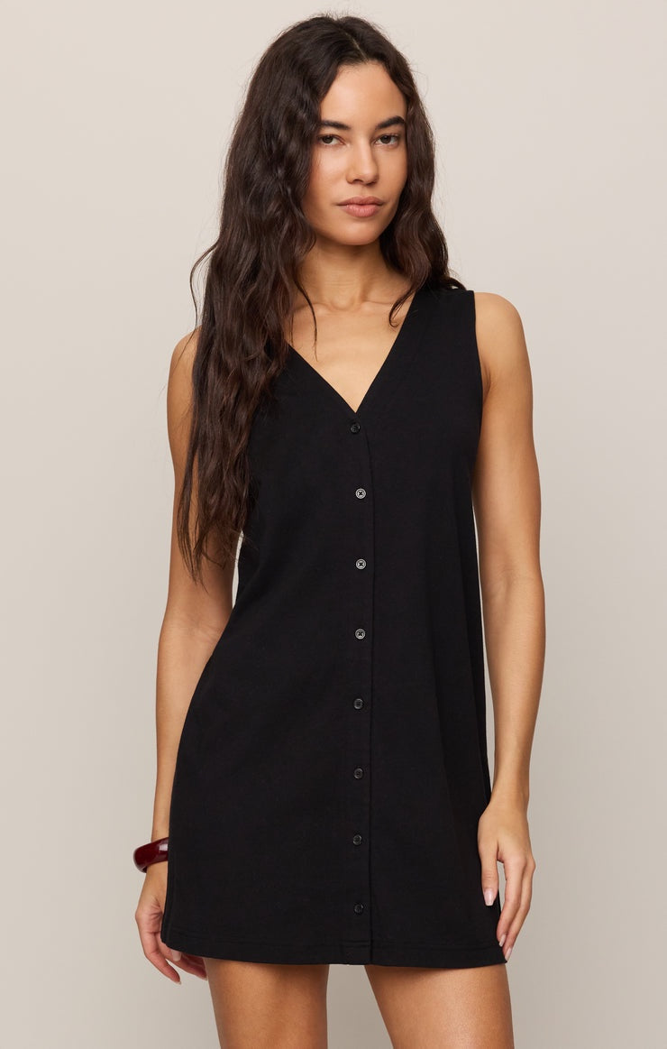 A woman with long, wavy brown hair models the Zertz SOLACE MINI DRESS TRUE BLACK—a sleeveless cotton mini dress with a V-neck and front buttons—against a plain light background.