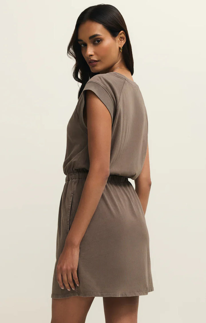 Woman with long dark hair models the Zertz KALANI DRESS PERFECTLY TAUPE—a short-sleeve, relaxed fit cotton jersey dress with an elastic waist—standing sideways and looking over her shoulder against a plain light background.