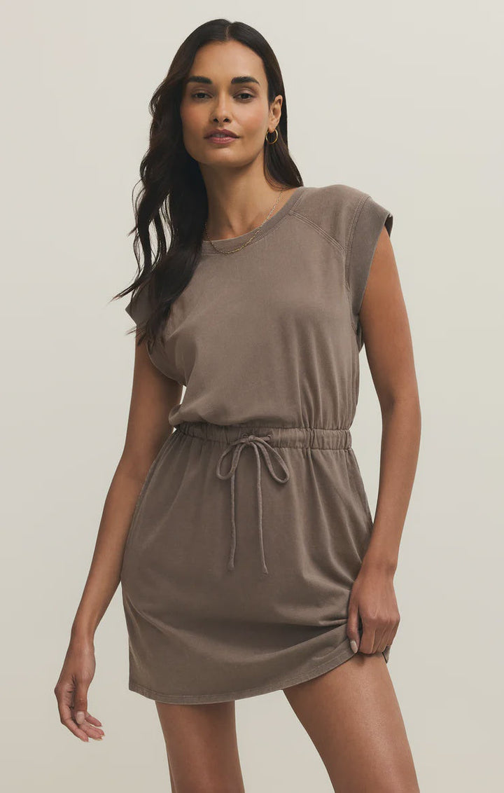 A woman models the Zertz KALANI DRESS PERFECTLY TAUPE—a short cotton jersey dress with cap sleeves and a drawstring waist—against a neutral background, posing with one hand on her hip.