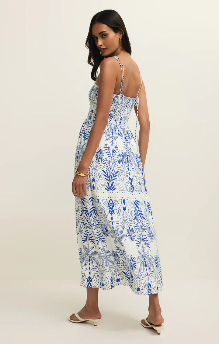 A woman models the Zertz NADINE PALM MAXI DRESS, featuring airy textured fabric with a botanical print, thin straps, and a smocked bodice. She pairs it with sandals and bracelets, glancing over her shoulder against a plain background.