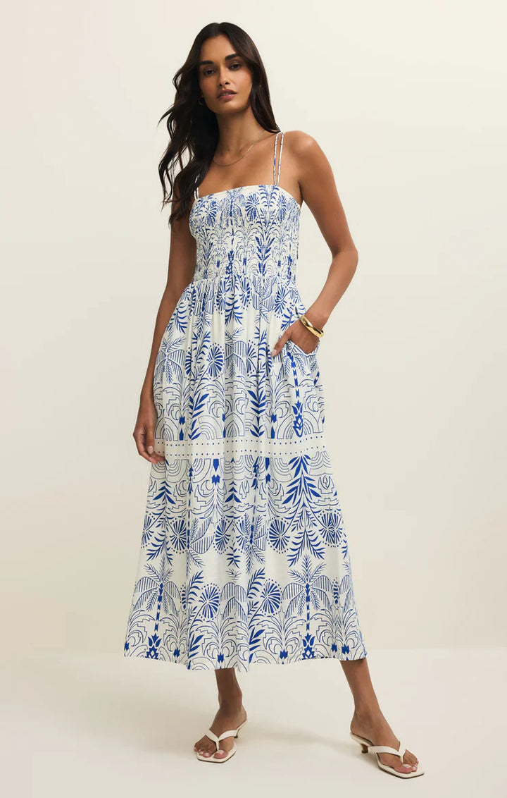 A woman with long dark hair poses against a plain backdrop wearing the Zertz NADINE PALM MAXI DRESS, a white and blue printed sundress with a smocked bodice and thin straps. She pairs it with sandals and casually places one hand in her pocket.