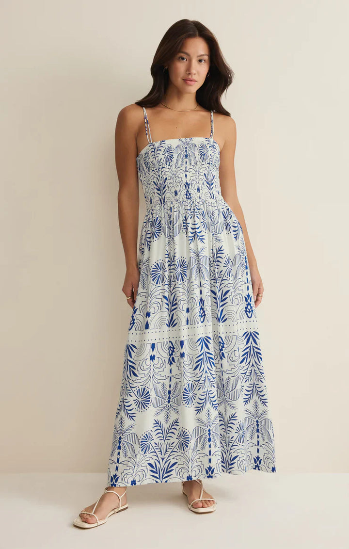 A woman stands against a plain wall wearing the Zertz NADINE PALM MAXI DRESS, a long sleeveless dress with blue botanical patterns and thin straps. Airy textured fabric and flat white sandals complete her fresh look.