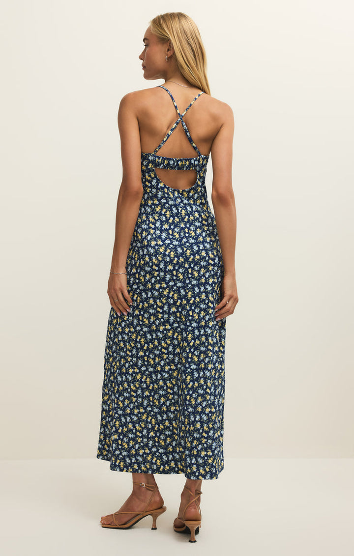 A woman faces away, wearing the Zertz CHIARA IDRA DITSY MIDI DRESS—sleeveless, blue floral with adjustable crisscross open-back straps—and strappy tan heels, standing against a plain off-white background.