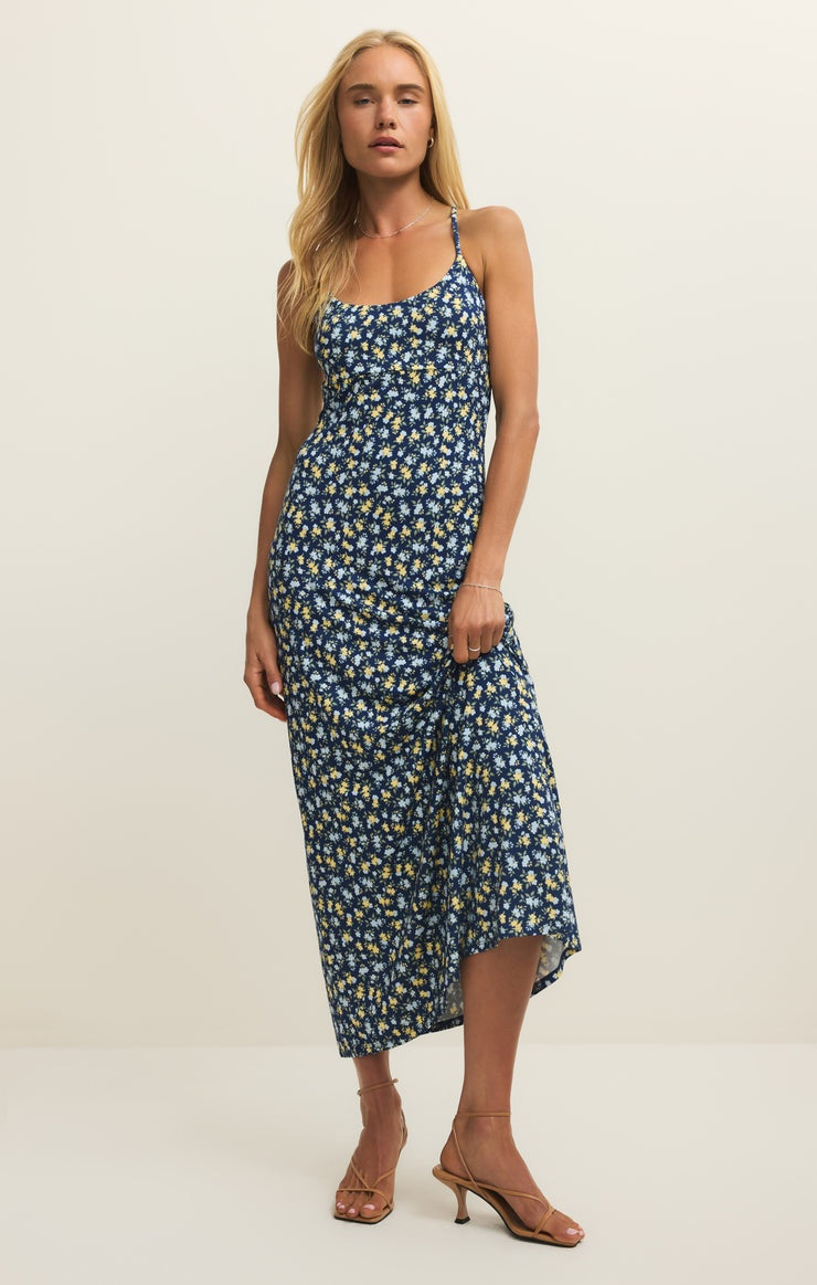 A woman with long blonde hair models the Zertz CHIARA IDRA DITSY MIDI DRESS—navy blue with white and yellow florals, adjustable straps—paired with tan heeled sandals, standing against a plain off-white background.