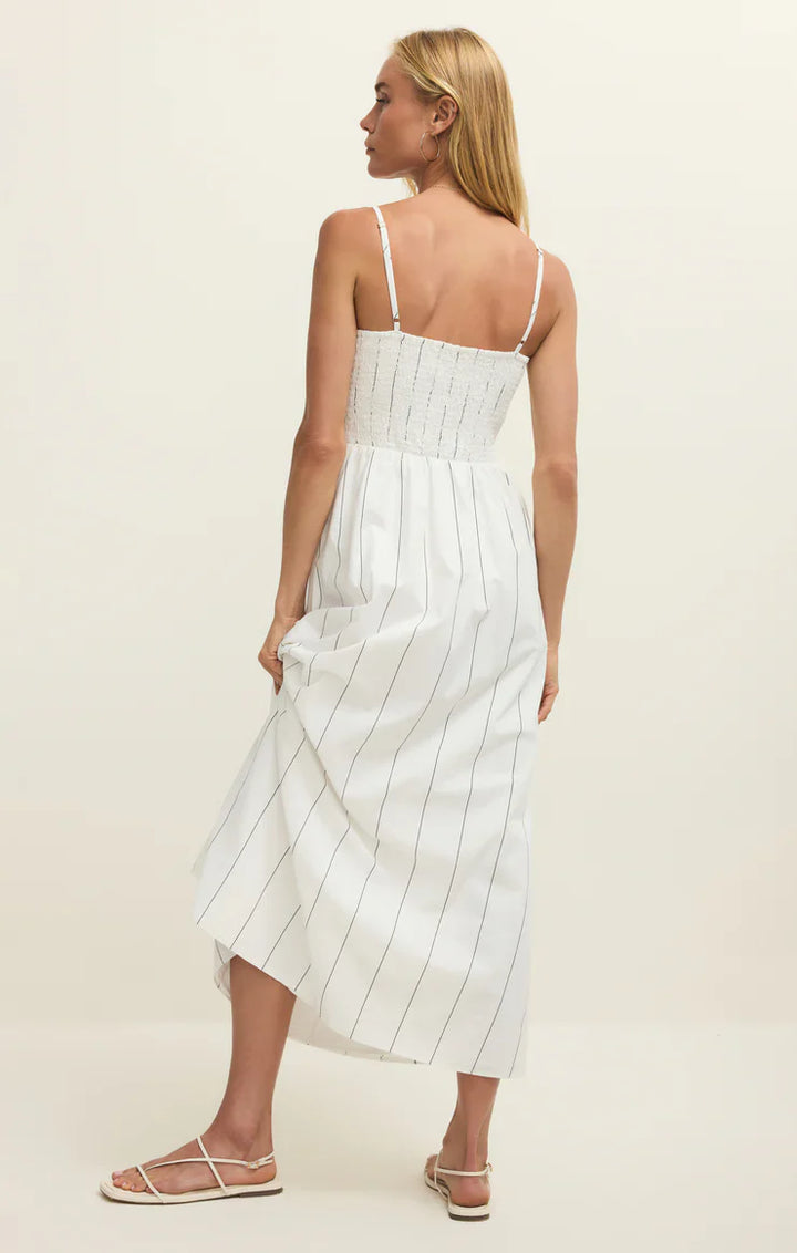 A woman with straight blonde hair and white sandals faces away, wearing the Zertz BAYSIDE STRIPE MIDI DRESS WHITE ZD261988—a white sleeveless dress with thin black stripes, smocked bodice, and thin straps.