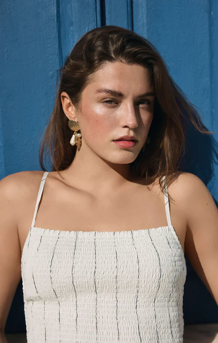 A woman with long brown hair wearing the Zertz BAYSIDE STRIPE MIDI DRESS WHITE ZD261988 and gold earrings stands before a bright blue wall, gazing to the side with a serious expression.