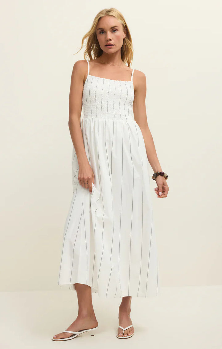 A woman wears the Zertz BAYSIDE STRIPE MIDI DRESS WHITE ZD261988, a sleeveless white dress with thin straps, black vertical pinstripes, and a smocked bodice. She pairs it with white sandals and a bracelet against a plain background.
