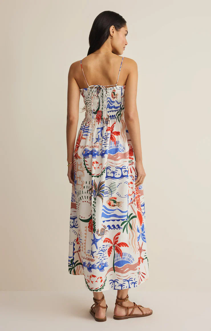 A woman faces away wearing the Zertz CORNELIA GETAWAY MIDI DRESS, a resort-ready coastal style with thin straps and sandals. The dress has a colorful red, blue, and green tropical print, smocked bodice, gathered back, and a neutral background.