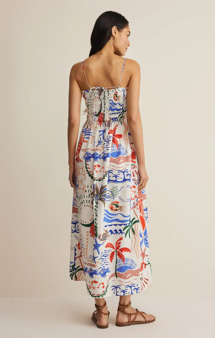 A woman faces away wearing the Zertz CORNELIA GETAWAY MIDI DRESS, a resort-ready coastal style with thin straps and sandals. The dress has a colorful red, blue, and green tropical print, smocked bodice, gathered back, and a neutral background.