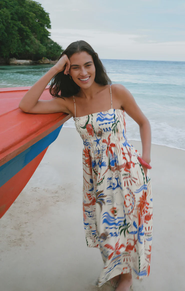 A woman wearing the Zertz CORNELIA GETAWAY MIDI DRESS, a colorful resort-ready sundress with a smocked bodice, smiles as she leans against a red and blue boat on a sandy beach with ocean and trees in the background.