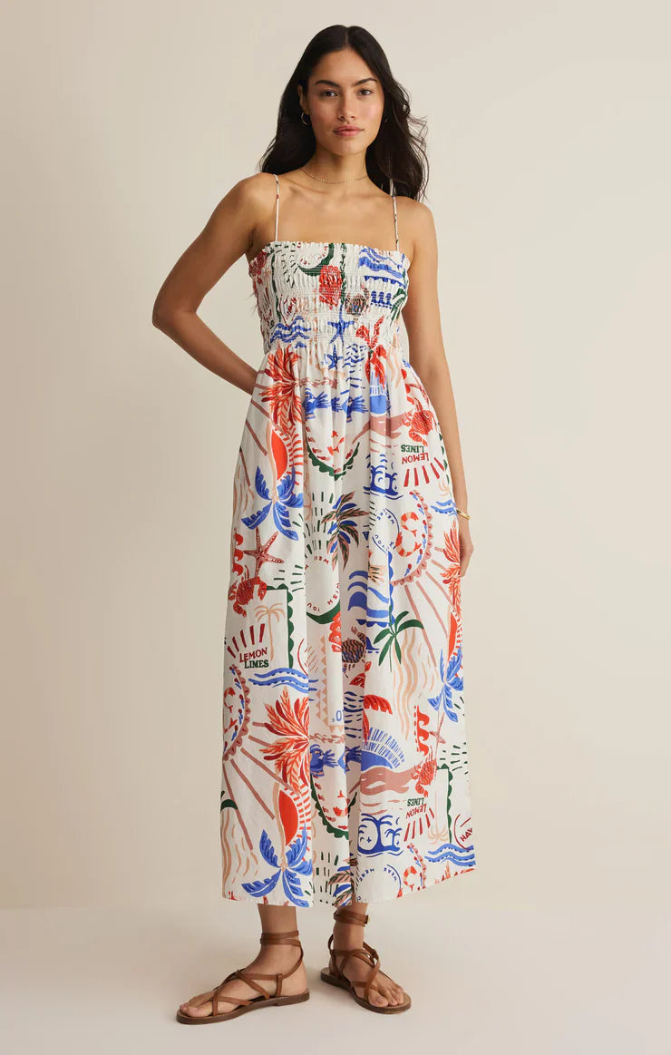 A woman stands against a plain beige background in the Zertz CORNELIA GETAWAY MIDI DRESS, a resort-ready, sleeveless dress with a smocked bodice and colorful abstract red, blue, and green patterns, paired with brown strappy sandals.