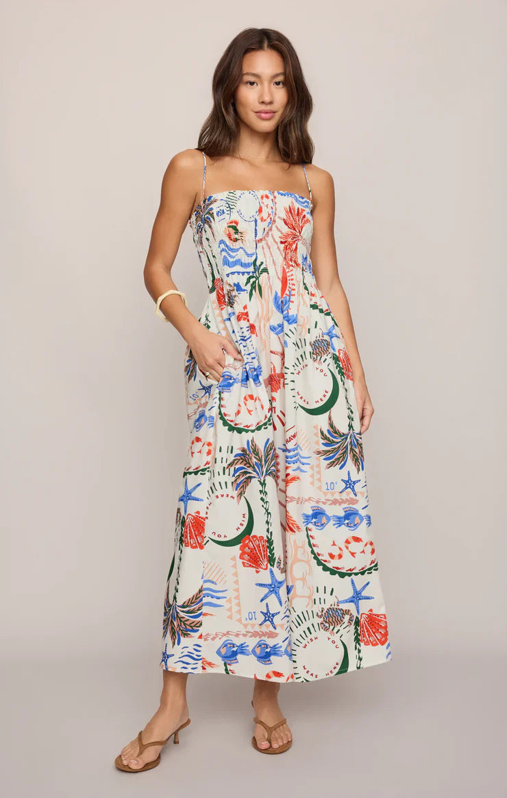 A woman wears the Zertz CORNELIA GETAWAY MIDI DRESS, a resort-ready, sleeveless dress with thin straps, smocked bodice, and vibrant tropical ocean-themed print. She has one hand in her pocket and pairs it with sandals.
