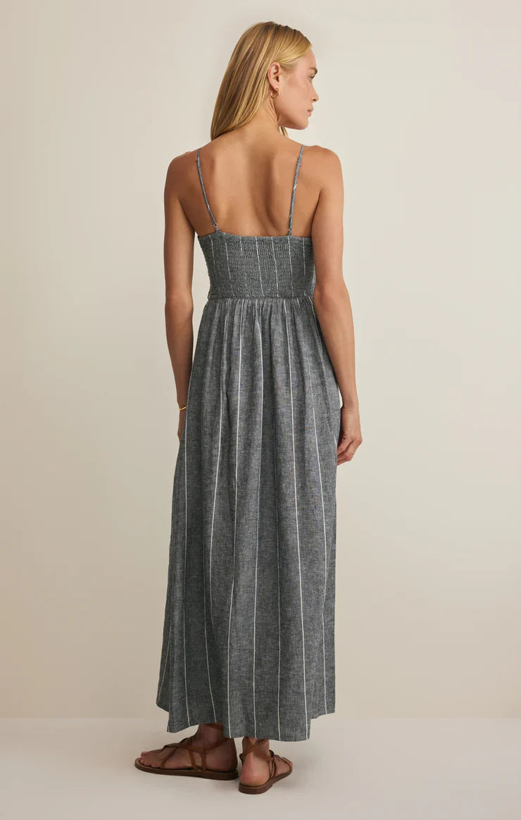A woman with long blonde hair faces away, wearing the Zertz ROMY STRIPED MAXI DRESS—a grey linen summer dress with a smocked bodice—and brown sandals, set against a plain light background.