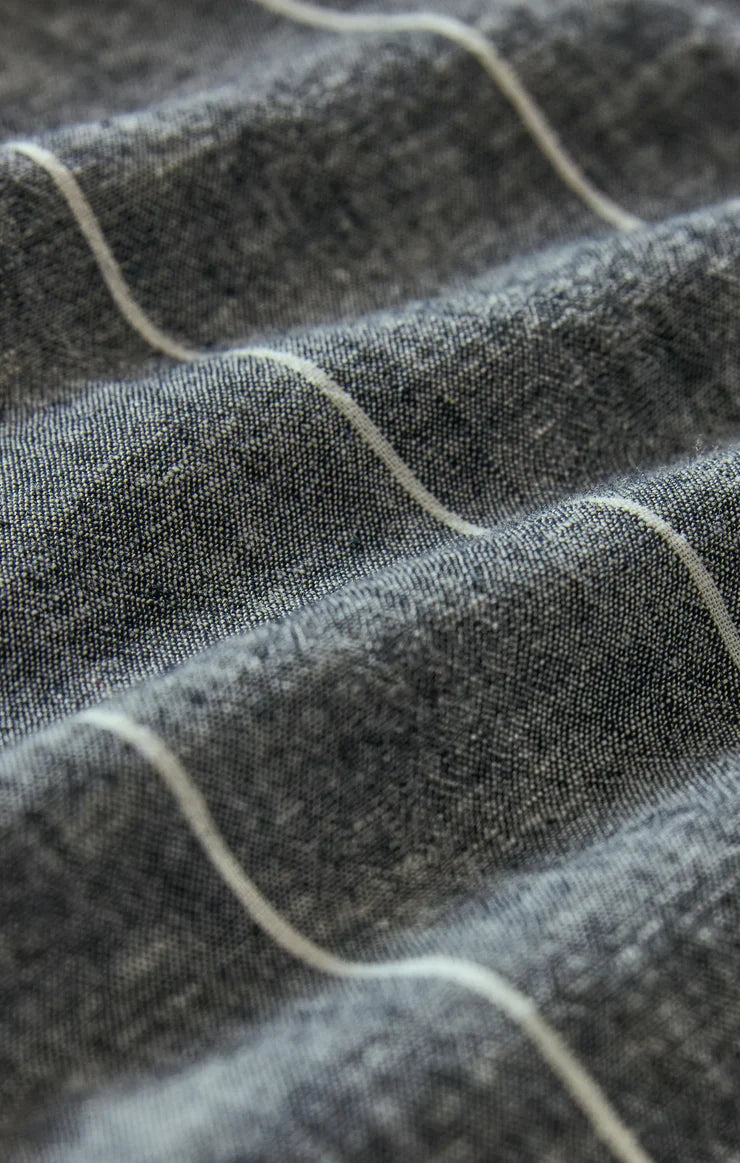 A close-up of the ROMY STRIPED MAXI DRESS by Zertz, showing textured dark gray fabric with thin white horizontal lines, evoking gentle waves and folds reminiscent of a linen summer dress.