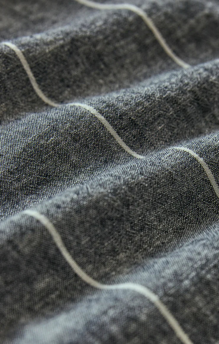 A close-up of the ROMY STRIPED MAXI DRESS by Zertz, showing textured dark gray fabric with thin white horizontal lines, evoking gentle waves and folds reminiscent of a linen summer dress.