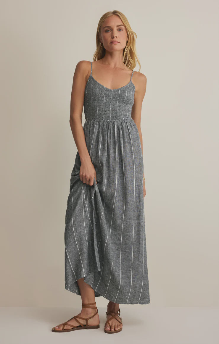 A woman stands against a plain background wearing the Zertz ROMY STRIPED MAXI DRESS, a blue-gray maxi dress with thin white stripes, paired with brown strappy sandals. She holds one side of the dress, highlighting its breezy style.