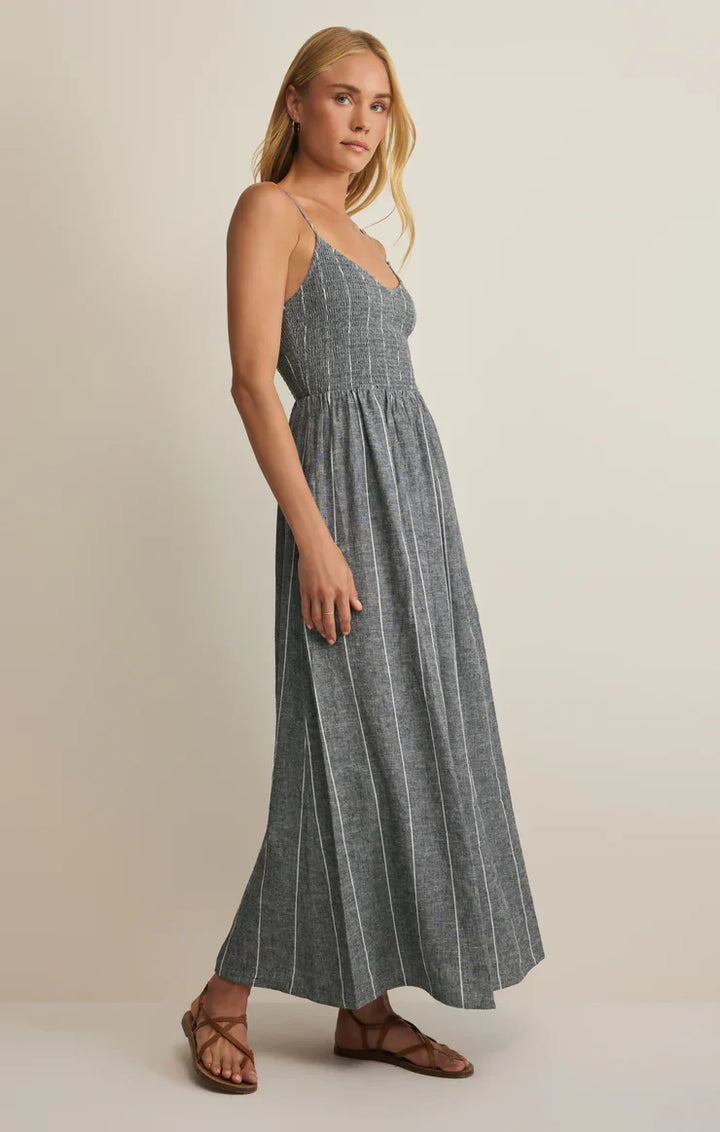 A woman stands sideways wearing the Zertz ROMY STRIPED MAXI DRESS—a blue-gray, striped linen maxi with a smocked bodice and thin straps—paired with brown strappy sandals, posing against a plain off-white background.